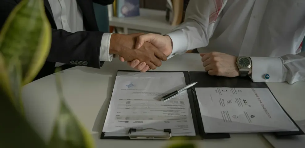 Two people shaking hands over a signed document folder and pen on a desk.